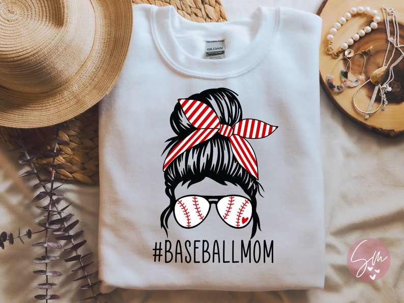 May include: White sweatshirt featuring a graphic of a woman with a messy bun wearing baseball-themed sunglasses and a red and white striped headband. The text "#BASEBALLMOM" is printed below the graphic.