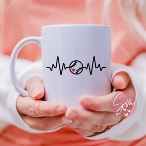 May include: White ceramic mug featuring a black and red baseball graphic and a heartbeat line. The mug is being held by a person's hands.