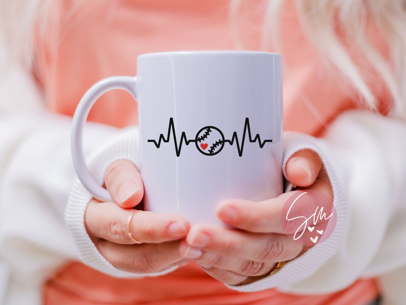 May include: White ceramic mug featuring a black and red baseball graphic and a heartbeat line. The mug is being held by a person's hands.