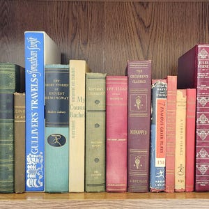 May include: A collection of vintage books on a wooden shelf. The spines display titles like "Gulliver's Travels" and "The Short Stories of Ernest Hemingway." Colors range from blue and green to red and tan, with decorative gold lettering.