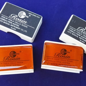 May include: Four containers of violin, viola, and cello rosin. Two containers are open and show the orange rosin inside. The containers are labeled "CLETO Rosin" and have the text "Violin-Viola-Cello Rosin Formula from Austria Leto Musical Instrument Co., Ltd. No 603"