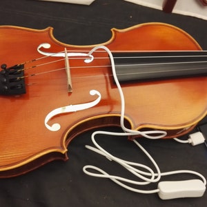 May include: A brown wooden violin with four strings and a white pickguard. A white cord connects the violin to a white power adapter and a black power bank.