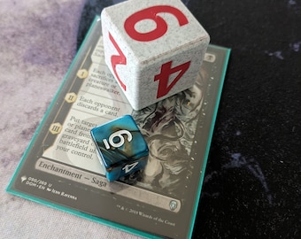 Puede incluir: Un dado azul y negro con el número 10, un dado blanco con los números 6 y 4, y una carta de Magic: The Gathering con el texto "Encantamiento - Saga" y "Cada oponente descarta una carta."