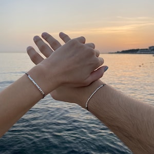 Könnte beinhalten: Zwei silberne Perlenarmbänder an schwarzen Kordeln. Die Armbänder werden an Handgelenken getragen, wobei die Hände aneinandergelegt sind. Der Hintergrund zeigt einen Sonnenuntergang über dem Meer mit Orange-, Gelb- und Blautönen.