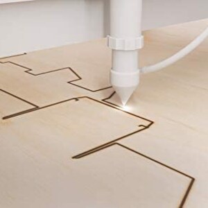 May include: A close-up of a laser cutter cutting a design into a piece of wood. The laser is white and the wood is light brown.