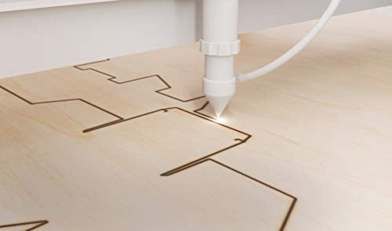 May include: A close-up of a laser cutter cutting a design into a piece of wood. The laser is white and the wood is light brown.