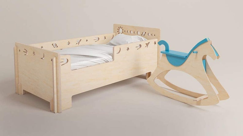 May include: A wooden toddler bed with a white sheet and a blue rocking horse. The bed has a decorative cutout design of stars, moons, and a sun.