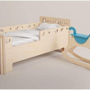 May include: A light wood toddler bed with a white sheet and a blue rocking horse. The bed has a decorative cutout design of stars, moons, and suns.