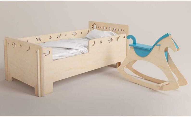 May include: A light wood toddler bed with a white sheet and a blue rocking horse. The bed has a decorative cutout design of stars, moons, and suns.