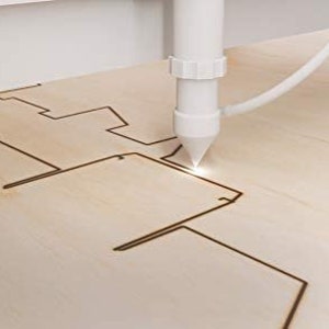 May include: A close-up of a laser cutter cutting a design into a piece of wood. The laser is white and the wood is light brown.