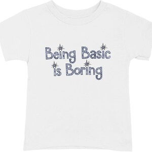 May include: A white short-sleeved t-shirt featuring the text "Being Basic is Boring" printed in a blue, dotted, bubble-style font. The design includes four small, dark blue starburst icons positioned around the lettering on the chest.