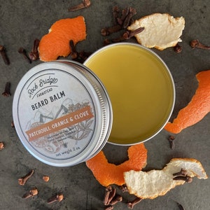 May include: A silver tin of Rock Bridge Farmstead beard balm with a label that reads "Beard Balm Patchouli, Orange & Clove" and "net weight 2 oz". The tin is open and the balm is visible inside. The tin is surrounded by orange peels and cloves.
