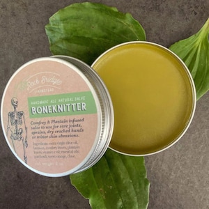 May include: A silver tin with a green label that reads "Roch Bridges Farmstead Handmade All Natural Salve Boneknitter Comfrey & Plantain infused salve to use for sore joints, sprains, dry cracked hands or minor skin abrasions. Ingredients: extra virgin olive oil, beeswax, comfrey leaves, plantain leaves, vitamin e oil; essential oils: patchouli, sweet orange, clove. Net weight 2 oz" The tin is open and the salve inside is a light yellow color.