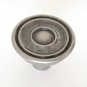 May include: A silver-toned metal knob with a circular design. The knob has a raised center and two concentric rings around it.