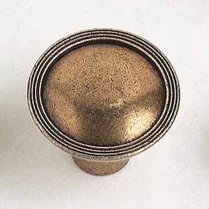 May include: A round, antique brass cabinet knob with a raised, textured center and three concentric rings around the edge.