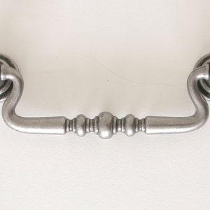 May include: A silver-toned cabinet pull with a decorative, rounded design. The pull has a curved shape and a center section with three raised, round accents.