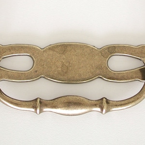 May include: A brass cabinet pull with a decorative, ornate design. The pull has a curved, oval-shaped handle and a rectangular plate with a cutout design.