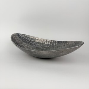 May include: An elongated, boat-shaped bowl with a dark, metallic finish. The interior surface features a distinct, raised grid pattern that resembles a textured reptile skin, while the exterior has a smooth, matte, dark gray appearance.