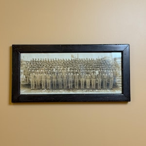 Puede incluir: Fotografía en blanco y negro enmarcada de un grupo numeroso de personas uniformadas. La fotografía está en un marco negro rectangular y cuelga de una pared beige. La fotografía parece ser un retrato de grupo.