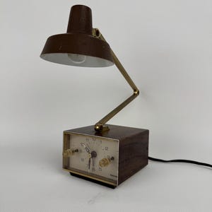 May include: A vintage brown desk lamp with a built-in clock. The lamp has a brown shade and a brass-colored adjustable arm. The clock face is white with black numerals and hands. The clock has two knobs and the word "benrus" is printed on the clock face.