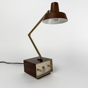 May include: A vintage brown desk lamp with a built-in clock. The lamp features a brown lampshade and a brass-colored adjustable arm. The clock face is white with black numerals and two clear knobs. The lamp is plugged into a black electrical cord.