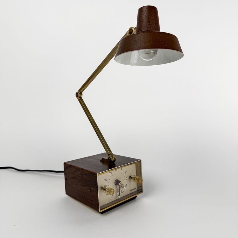 May include: A vintage brown desk lamp with a built-in clock. The lamp features a brown lampshade and a brass-colored adjustable arm. The clock face is white with black numerals and two clear knobs. The lamp is plugged into a black electrical cord.