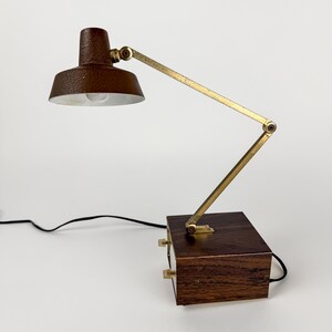 May include: A vintage desk lamp with a brown, textured shade and a brass-colored adjustable arm. The lamp sits on a dark wooden base with two knobs. A black power cord extends from the base.