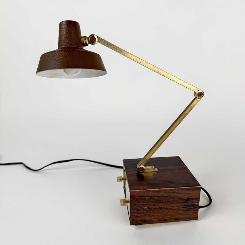 May include: A vintage desk lamp with a brown, textured shade and a brass-colored adjustable arm. The lamp sits on a dark wooden base with two knobs. A black power cord extends from the base.