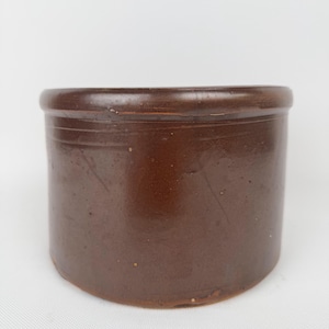 May include: A brown, cylindrical stoneware crock with a rounded rim. The antique jar has a smooth, glossy finish and a slightly tapered base. The crock is likely used for food storage and has a rustic, vintage aesthetic.