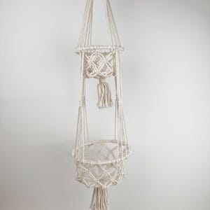 Puede incluir: Colgador de plantas de macramé color crema con dos cestas tejidas y una borla. El soporte para plantas está suspendido de un solo cordón y presenta intrincados patrones de nudos. Las cestas están diseñadas para contener plantas pequeñas y medianas.