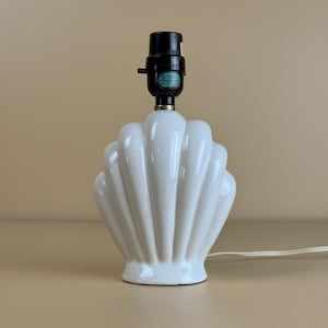 May include: A white ceramic table lamp base shaped like a scallop shell with a glossy finish. The lamp features a black socket at the top with a small switch and a white electrical cord extending from the base. The design is symmetrical and ribbed.