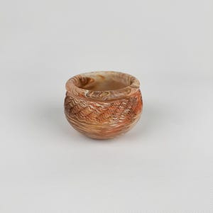 May include: A small, round, decorative bowl with a textured surface. The bowl features a blend of brown, orange, and cream colors, creating a marbled effect. The rim is smooth, while the body has a carved, patterned design.