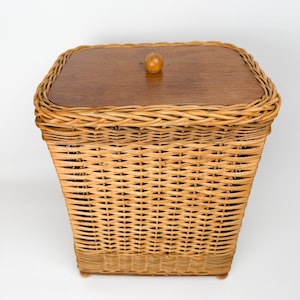 May include: A rectangular wicker basket with a wooden lid and a small, round knob. The basket is woven with light brown wicker and has a slightly tapered shape. The lid is a darker brown wood.