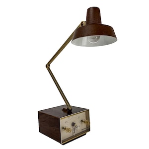 May include: A vintage brown desk lamp with a built-in clock. The lamp features a brown lampshade and a brass-colored adjustable arm. The clock face is white with black numerals and two clear knobs. The lamp is on a dark brown wooden base.