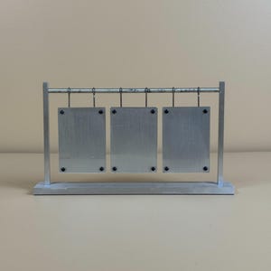 Könnte beinhalten: Ein silberner Metall-Ausstellungsständer mit drei rechteckigen Tafeln, die an einer horizontalen Stange hängen. Jede Tafel ist mit schwarzen Schrauben befestigt. Der Ständer hat eine rechteckige Basis und vertikale Stützen.