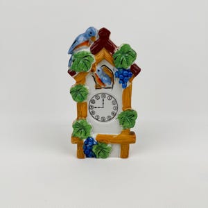May include: A miniature cuckoo clock with a white face and a wooden frame. The clock features a small blue bird on top and another inside the clock face. Green leaves and blue grapes decorate the frame. The clock hands point to 10:09.