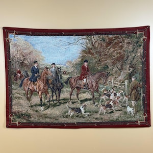 May include: A rectangular woven wall tapestry featuring a fox hunting scene with riders on horseback and a pack of hounds in a wooded landscape. The border is a deep red fabric with a repeating black horse bit and bridle pattern throughout.