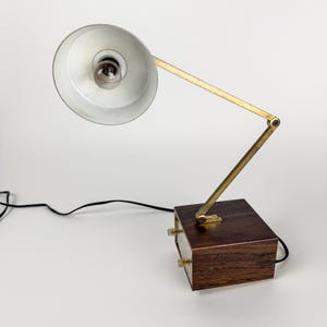 May include: A vintage desk lamp with a white lampshade and a brass-colored adjustable arm. The lamp sits on a dark wood base with two small brass knobs. A black power cord extends from the base.