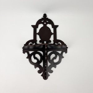 May include: Dark brown wooden wall shelf with an ornate, gothic-inspired design. The shelf features a decorative top with a central oval shape and a lower shelf with scrolled details. The shelf is mounted on a white wall.