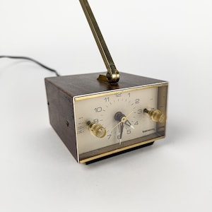 May include: A vintage brown wooden alarm clock with a gold-colored frame and a gold-colored adjustable arm. The clock face is white with black numbers and gold-colored hands. The clock has two clear knobs and a black power cord.