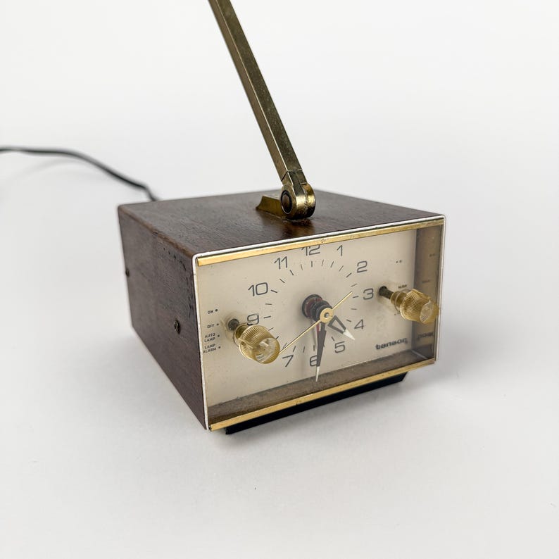 May include: A vintage brown wooden alarm clock with a gold-colored frame and a gold-colored adjustable arm. The clock face is white with black numbers and gold-colored hands. The clock has two clear knobs and a black power cord.