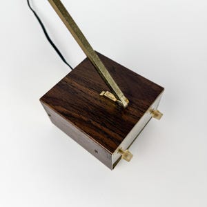 May include: A vintage desk lamp with a dark wood base and a brass arm. The base is rectangular with two small knobs on the front. The brass arm extends upward and away from the base. A black cord is attached.