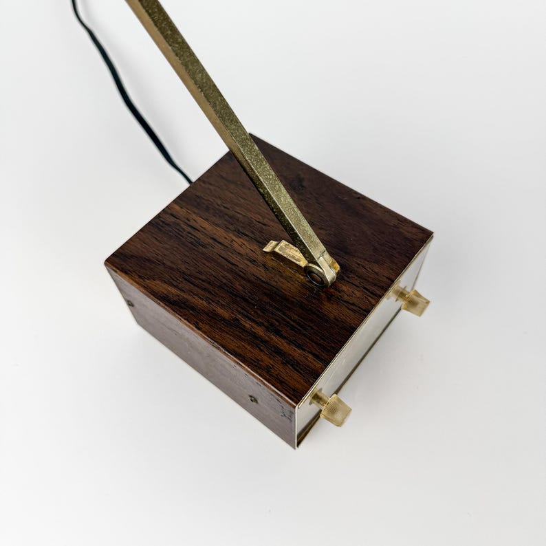 May include: A vintage desk lamp with a dark wood base and a brass arm. The base is rectangular with two small knobs on the front. The brass arm extends upward and away from the base. A black cord is attached.
