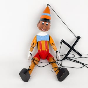 Könnte beinhalten: Eine Holzmarionette von Pinocchio mit einem orangefarbenen Hut, Hemd und roter Hose. Die Marionette hat eine blaue Fliege, schwarze Schuhe und weiße Handschuhe. Schwarze Schnüre sind zur Manipulation befestigt.