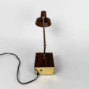 May include: A vintage desk lamp with a dark brown shade and a brass-colored adjustable arm. The lamp sits on a rectangular wooden base with a brass-colored front panel. A black power cord extends from the base.