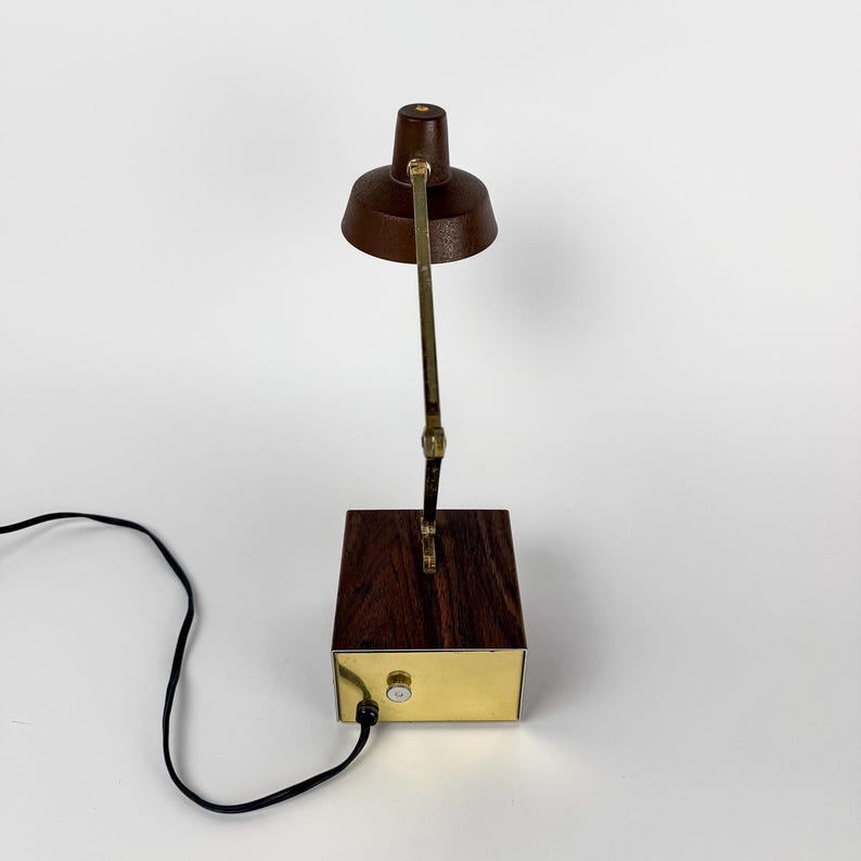 May include: A vintage desk lamp with a dark brown shade and a brass-colored adjustable arm. The lamp sits on a rectangular wooden base with a brass-colored front panel. A black power cord extends from the base.