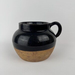 May include: A small, handcrafted ceramic mug with a rounded body and a handle. The top half is glazed in glossy black, contrasting with the unglazed, tan-colored bottom half. The mug is designed for beverages.
