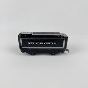 May include: A black toy train car with the words "NEW YORK CENTRAL" in white lettering. The train car has a rectangular shape with wheels and a detailed roof. The overall design is vintage.