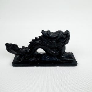 Könnte beinhalten: Eine schwarze Drachenfigur mit aufwendigen Details ruht auf einem rechteckigen Sockel. Der Drache ist in einer dynamischen Pose dargestellt, mit erhobenem Kopf und gewundenem Körper. Die Figur besteht aus einem dunklen, möglicherweise polierten Material.