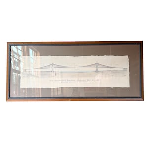 May include: A framed architectural print of the Brooklyn Bridge featuring a detailed line drawing on off-white paper with deckled edges. The text reads: THE BROOKLYN BRIDGE - OPENED MAY 24, 1883. The print is housed in a dark wood frame with a black border.